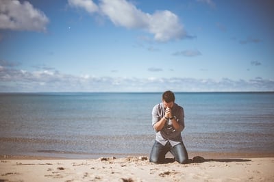 A man kneeling on the beach facing away from the water with his hands folded in prayer with his head bowed and resting on this hands - typically known as a prayer position