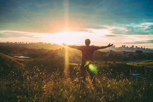 A man standing in a field on top of a hill overlooking a city during sunrise