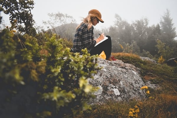 woman sitting on a rock surrounded by nature journaling.